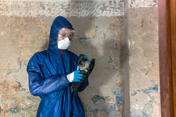 Trabalhador em terno de proteção azul, óculos e respirador segurando um moedor de ângulo
 - Foto, Imagem