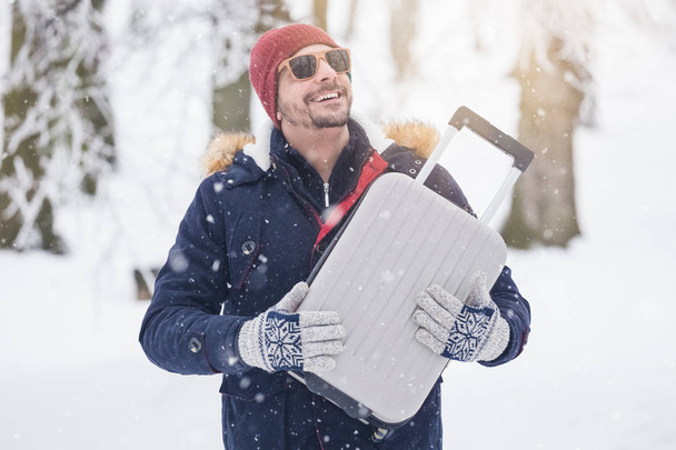 Portrait of young fashionable man on snow holding suitcase. Winter vacation travel concept. - Photo, Image