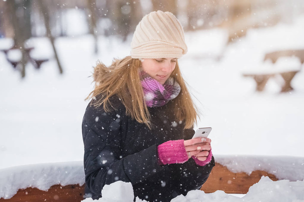 Beautiful woman sitting on bench in the park and using smart phone winter landscape. - Photo, Image