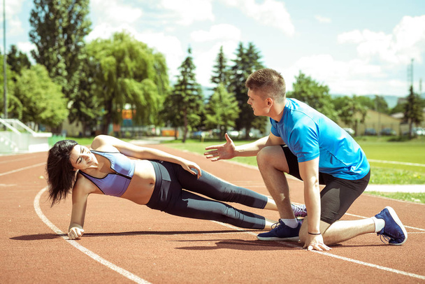 Beautiful brunette woman doing side plank exercise outdoors with support of her personal trainer. Toned image. - Фото, зображення