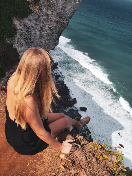 Top view of blonde woman tourist sitting on the cliff and admire beautiful ocean view - Photo, image
