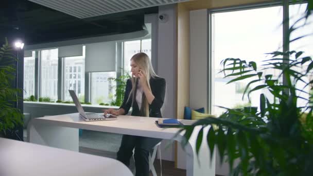 Trendy zakenvrouw die laptop kijkt en telefoneert. Stijlvolle jonge blonde vrouw werkt in het kantoor aan tafel surfen laptop en het hebben van een telefoontje terwijl glimlachen. - Video