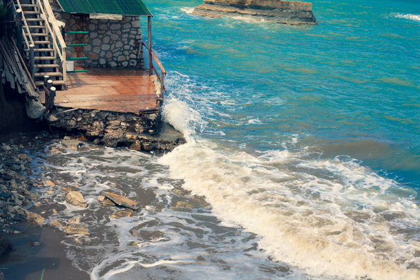 Wellen krachen gegen Felsen. Ozeanbewegungen gegen die Steine. Blaues Meer, das ein zerstörtes Haus zerstört. Die mediterrane Brandung zertrümmert Holztreppen. Flüssig zuschlagende Steinruinen. - Foto, Bild