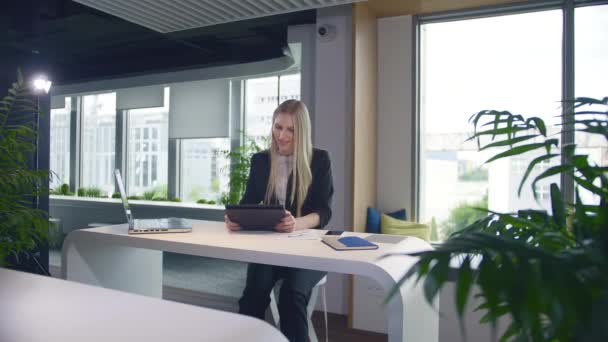 Stijlvolle zakenvrouw met tablet op kantoor. Elegante vrouw in pak zittend aan tafel met laptop en surftablet in modern lichtkantoor met grote ramen. - Video