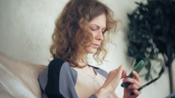 Young beautiful business woman sitting on sofa at table with tablet and using smartphone - Záběry, video