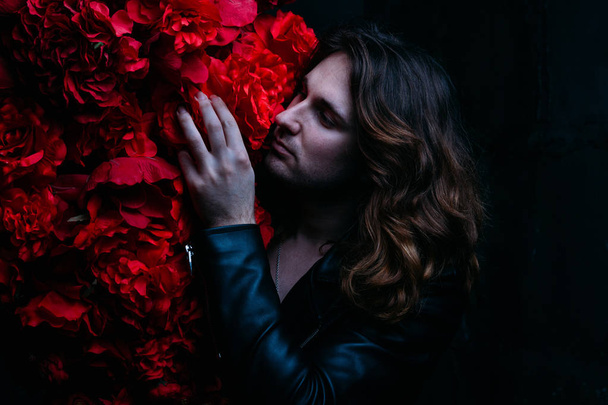 brutal man in a leather jacket on a naked body with long hair stands near the decor with red flowers flowers. strength and beauty. protect beauty. hair care. - Zdjęcie, obraz