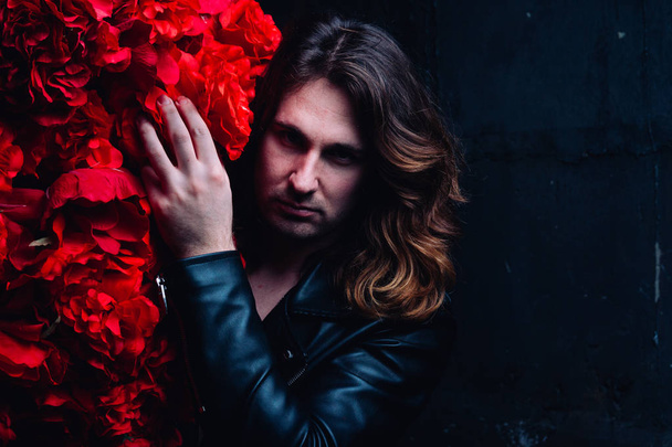 brutal man in a leather jacket on a naked body with long hair stands near the decor with red flowers flowers. strength and beauty. protect beauty. hair care. - Photo, image