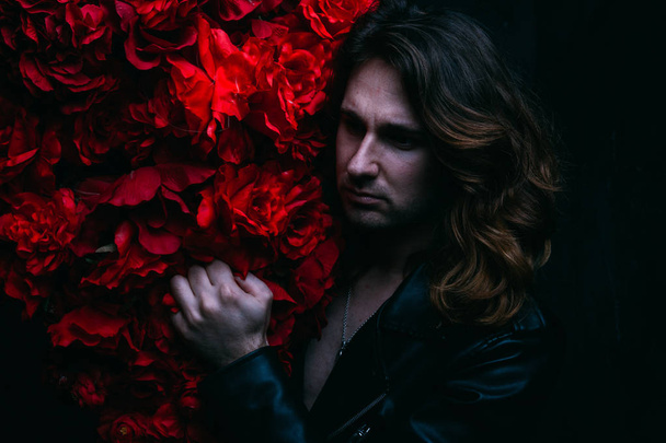 brutal man in a leather jacket on a naked body with long hair stands near the decor with red flowers flowers. a lonely man is sad and desperate - 写真・画像
