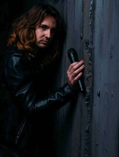 brutal man in a leather jacket on a naked body with long hair. holds a microphone in his hand, leans against the wall. singer - Фото, зображення