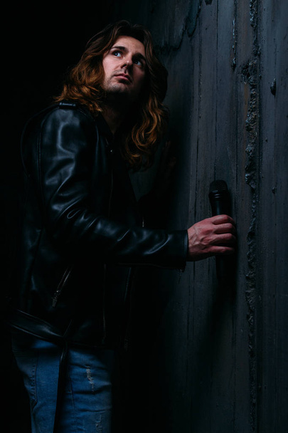 brutal man in a leather jacket on a naked body with long hair. holds a microphone in his hand, leans against the wall. singer. looks back - Fotoğraf, Görsel