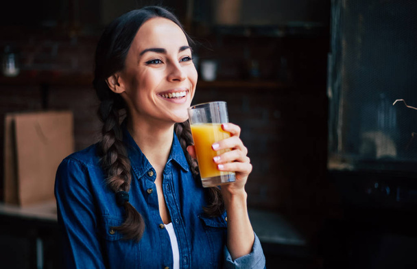 Retrato de una joven morena muy guapa con camisa vaquera en una cocina en casa. Chica riendo y sosteniendo jugo de naranja en un vaso transparente
. - Foto, imagen