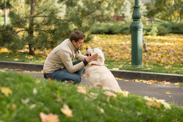    Młody człowiek sobie kurtkę beż i dżinsy jest przyjazny z jego duży pies Labrador w jesienny park. Patrzą na siebie, oni mają wzajemne zrozumienie. - Zdjęcie, obraz