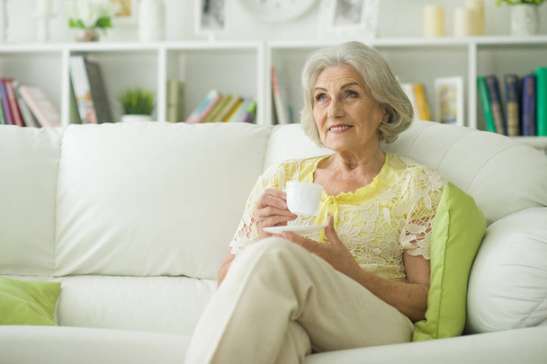 Beautiful senior woman sitting and drinking tea on sofa at home - Фото, изображение