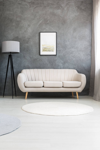 Poster on a gray wall, above a beige, leather sofa and a tripod floor lamp in a modern living room interior - Foto, Imagen