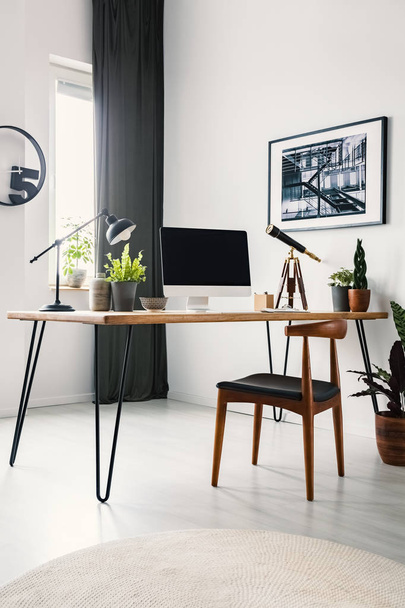 Wooden chair standing by the hairpin desk with empty mockup monitor, lamp and fresh potted plants standing in bright living room interior with poster on the wall and window with drape - 写真・画像