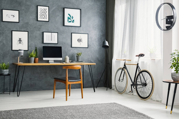 Bicycle next to wooden chair at desk in grey home office interior with posters. Real photo - 写真・画像