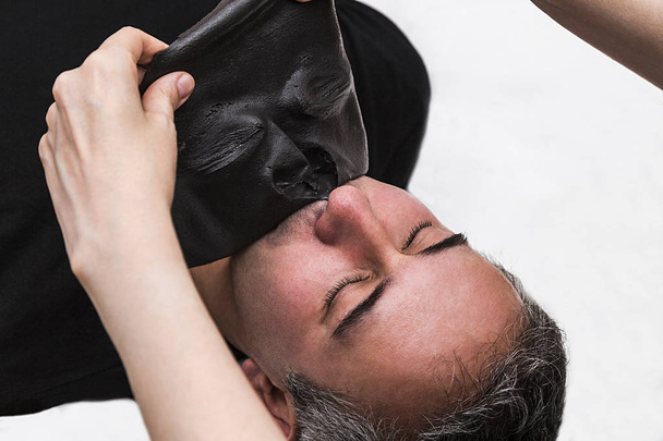 Лікар видаляє шкіру алкінату морських водоростей чорну маску з обличчя пацієнта чоловічої статі
 - Фото, зображення