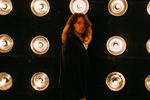 portrait a man in a black jacket and in a black shirt, with long hair, stands against the backdrop of bright lanterns, safites. on the stage. performance.turn around - Фото, зображення