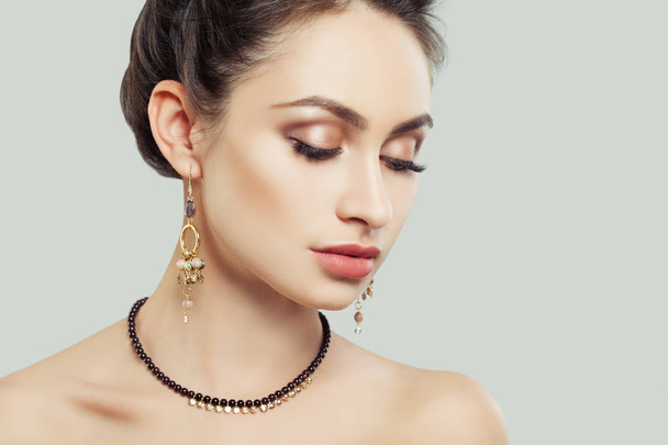Перфектная девушка с макияжем и украшениями на фоне белой стены. Золотые серьги и ожерелье с полудрагоценными камнями
 - Фото, изображение