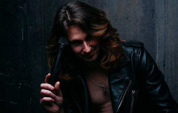 brutal man in a leather jacket on a naked body with long hair. a lonely man is sad and desperate. holds a microphone in his hand, leans against the wall.smiles - Foto, Bild
