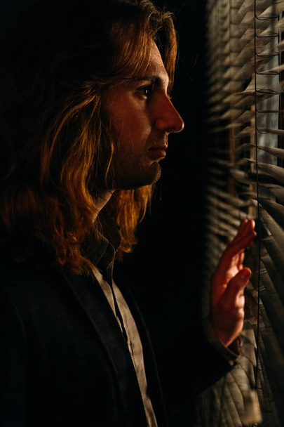 a man with long hair is standing near the window, opening the blinds with lobes,in a black jacket. look at the street, spy on people,sunrise. looking out the window - Foto, Imagen