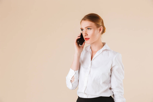 Photo of serious young business woman standing isolated over beige wall backgound looking aside talking by mobile phone. - Photo, image
