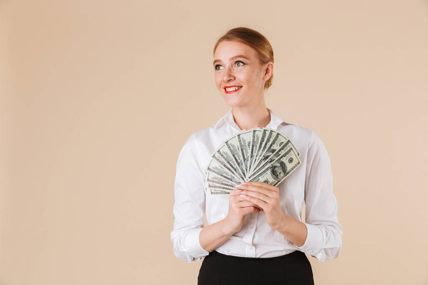 Portrait of a smiling businesswoman showing money banknotes isolated over beige background - Fotó, kép