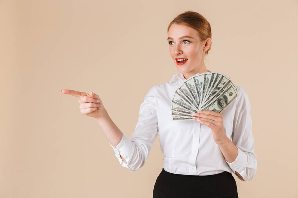 Portrait of a smiling businesswoman showing money banknotes and pointing finger at copy space isolated over beige background - Foto, Imagem