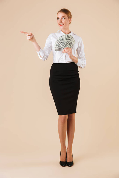 Full length portrait of a smiling businesswoman showing money banknotes and pointing finger at copy space isolated over beige background - Foto, imagen
