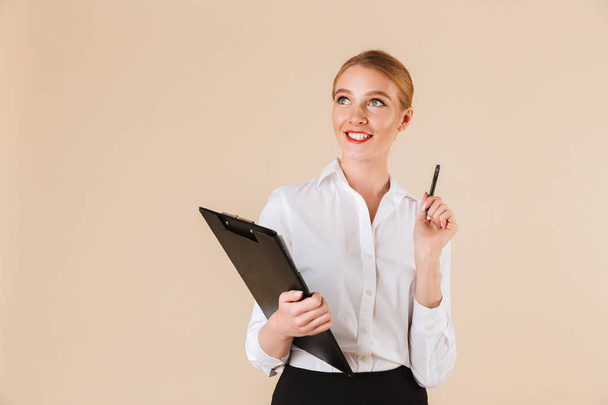 Image of young happy business woman isolated over beige wall backgound holding clipboard. Looking aside. - Fotoğraf, Görsel