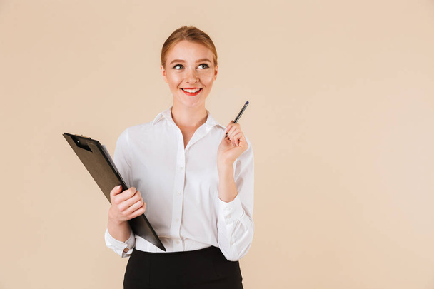 Image of young happy business woman isolated over beige wall backgound holding clipboard. Looking aside pointing. - Fotografie, Obrázek