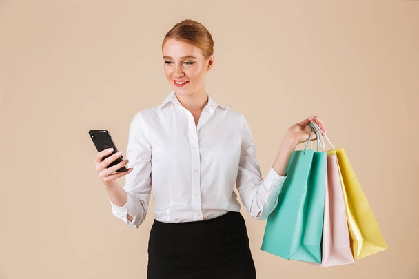 Image of cheerful young business woman standing isolated over beige wall backgound looking aside holding shopping bags using mobile phone. - 写真・画像