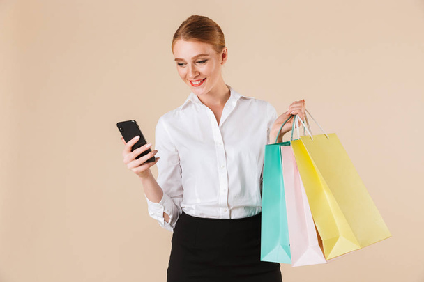 Image of cheerful young business woman standing isolated over beige wall backgound looking aside holding shopping bags using mobile phone. - Fotografie, Obrázek