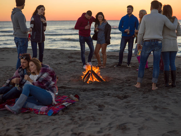 Young Couple Sitting with friends Around Campfire on The Beach At sunset drinking beer - Fotó, kép