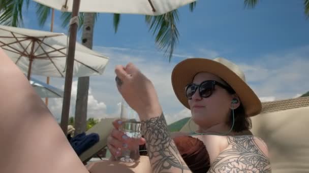 Woman in summer hat lying on sunbed and drinking water by the pool - Imágenes, Vídeo