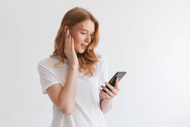 Photo of happy young redhead woman isolated over white wall background listening music with earphones by mobile phone. - 写真・画像