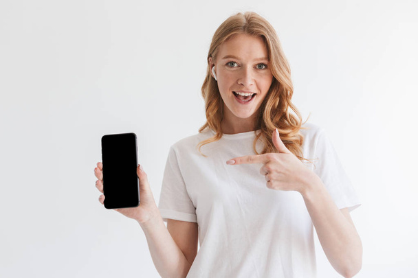 Photo of happy young redhead woman isolated over white wall background showing display of mobile phone pointing listening music. - Foto, immagini
