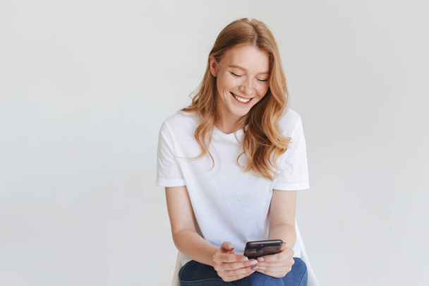 Image of young redhead happy woman isolated over white wall background using mobile phone. - 写真・画像