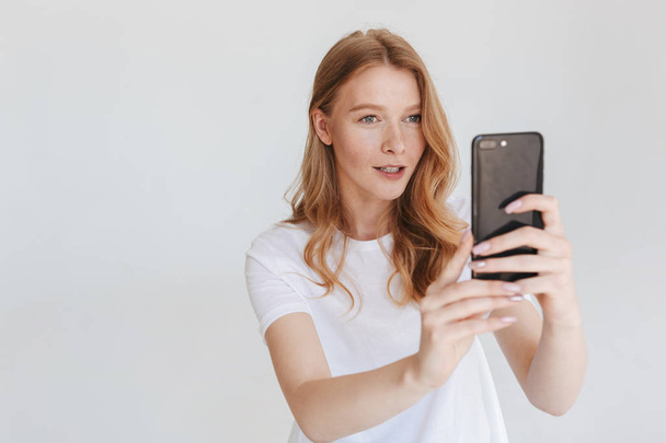 Image of young redhead woman isolated over white wall background using mobile phone make photo. - 写真・画像