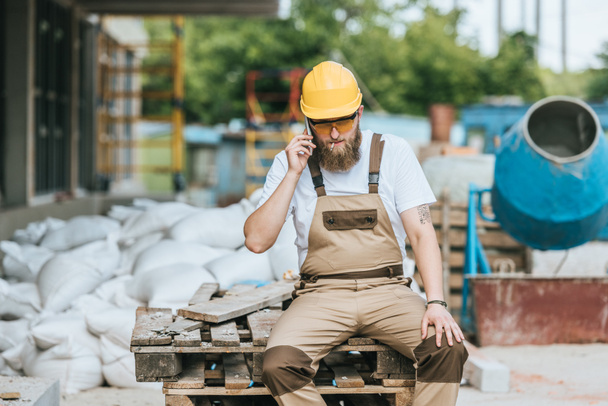 builder in hardhat and protective googles smoking cigarette and talking on smartphone at construction site  - Photo, Image
