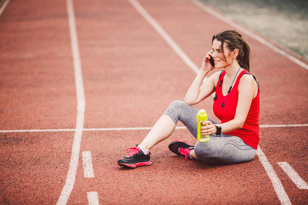 Sport and technology. A beautiful young Caucasian woman with ponytail sitting resting after workout during run at stadium, a red treadmill track. Uses for cell phone headphones and sports watches. - Φωτογραφία, εικόνα