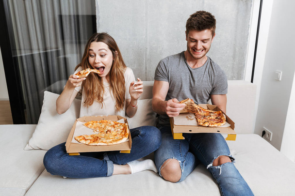 Happy young couple eating pizza while sitting together on couch at home - Фото, зображення