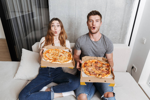 Excited young couple holding boxes with pizza while sitting together on couch at home - Foto, Imagen