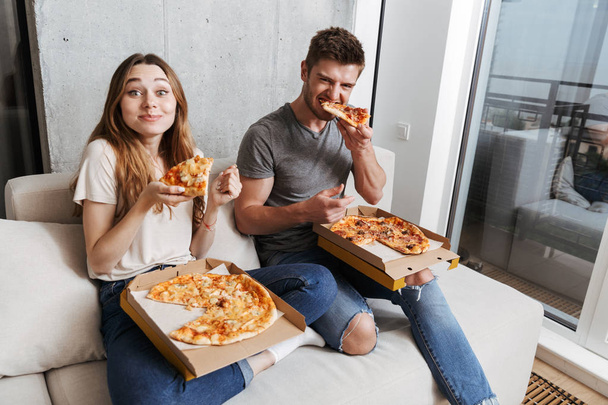 Satisfied young couple eating pizza while sitting together on couch at home - Photo, Image