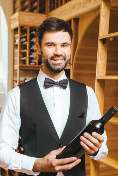sonriente joven administrador de vinos con botella de vino de lujo en la tienda de vinos
 - Foto, Imagen