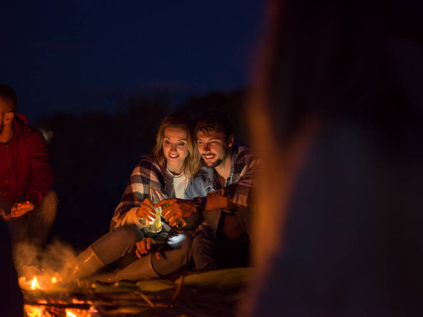 Young Couple Sitting with friends Around Campfire on The Beach At Night drinking beer - Foto, afbeelding