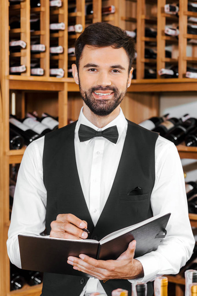 handsome young wine steward making notes and looking at camera at wine store - Foto, Bild