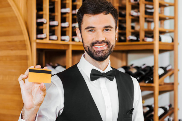 smiling young wine steward holding golden card at wine store - Fotoğraf, Görsel