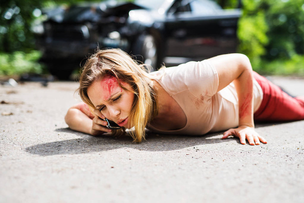 Mujer joven lesionada yaciendo en la carretera después de un accidente de coche, haciendo una llamada telefónica
. - Foto, Imagen