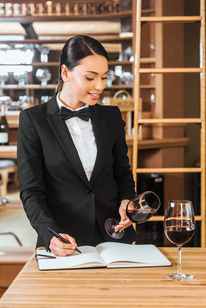 beautiful female wine steward looking at glass of wine and writing in notebook at wine store - Φωτογραφία, εικόνα